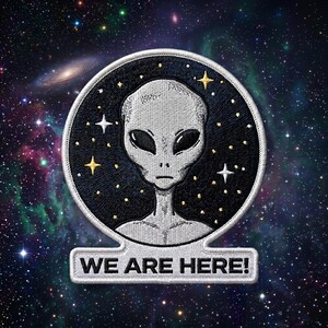 May include: A circular embroidered patch featuring a grey alien head centered against a black background filled with small gold and white stars. Below the circle, a rectangular tab displays the white embroidered text WE ARE HERE! in capital letters.