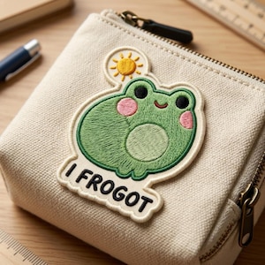 May include: A beige canvas pouch with a zipper closure, featuring an embroidered patch of a green frog with pink cheeks and the text "I FROGOT". A yellow sun is above the frog's head.