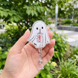 May include: A clear acrylic badge reel featuring a white ghost illustration holding a silver Diet Coke can with a red and white striped straw. The ghost has black eyes and a small mouth, with a retractable cord and a metal clip on the back.