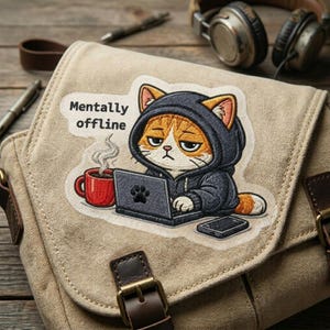 May include: A beige canvas messenger bag with a patch featuring a cartoon cat wearing a hoodie and using a laptop. The patch includes the text "Mentally offline" and a red coffee mug. A smartphone is also visible.