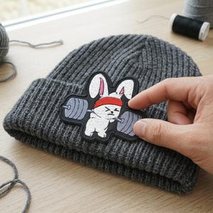 May include: A gray ribbed knit beanie with a cartoon patch of a white bunny lifting weights. The bunny wears a red headband. The patch is black with white, gray, and red embroidery.