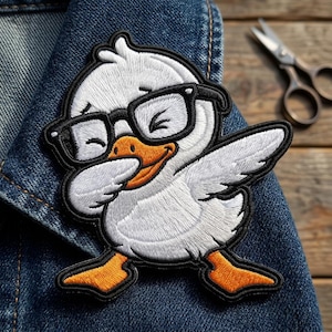 May include: Embroidered patch featuring a white duck wearing black-framed glasses, dabbing with its wing. The duck has an orange beak and feet, with black outlines. The patch is on a denim jacket.