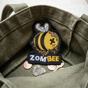 May include: A canvas tote bag in olive green holds a Zombee patch, featuring a yellow and black bee with a zombie brain and a cross for an eye. The patch is on a black background with the word "ZOMBEE".