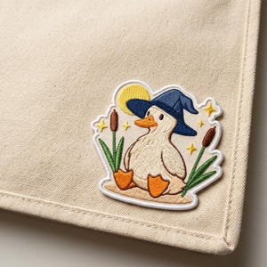 May include: Embroidered patch featuring a cartoon duck wearing a blue witch hat, sitting on a sandy area with cattails and grass. A yellow moon and stars are in the background. The patch is on a beige fabric.