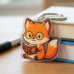 May include: A clear acrylic keychain featuring a cartoon fox with orange fur, wearing green glasses, and reading a brown book. The fox has a white-tipped tail and is attached to a silver keyring. The keychain is a cute accessory.