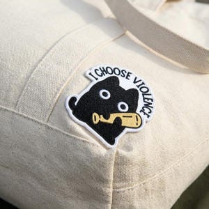 May include: Cream-colored canvas tote bag with a black cat patch. The patch features a black cat holding a yellow object, with the text "I CHOOSE VIOLENCE" in white.