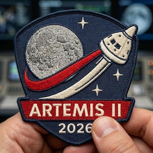 May include: Embroidered patch featuring a stylized depiction of the moon, a spacecraft, and the text "ARTEMIS II 2026." The patch has a navy blue background with red, white, and gold accents. Four stars are embroidered around the spacecraft.