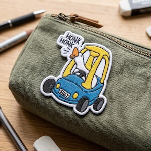 May include: A green canvas pencil case with a patch featuring a cartoon goose driving a blue car. The patch has the word "SILLY" and speech bubbles saying "HONK HONK". Other items, including pens and an eraser, are visible.