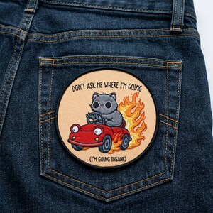 May include: Embroidered patch featuring a gray cat driving a red car engulfed in flames. The patch has the text "DON'T ASK ME WHERE I'M GOING" and "(I'M GOING INSANE)" on a tan background, sewn onto a blue denim pocket.