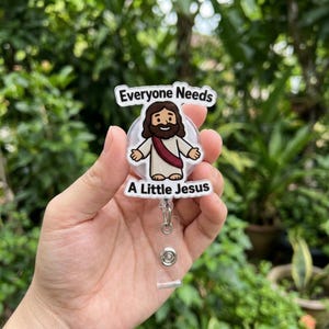 May include: A clear acrylic badge reel featuring a cartoon illustration of Jesus with brown hair and a beard, wearing a white robe with a red sash. The text "Everyone Needs A Little Jesus" is printed in black letters above and below the figure.