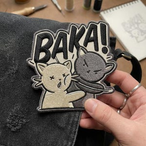 May include: Embroidered patch featuring two cartoon cats, one slapping the other. The patch has the word "Baka!" in bold letters. The cats are in shades of gray and beige, with black outlines. The patch is on a black fabric.