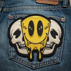 May include: Embroidered patch featuring a yellow smiley face with melting details and two white skulls on a blue denim background. The smiley face has black eyes with star accents and a dripping design.