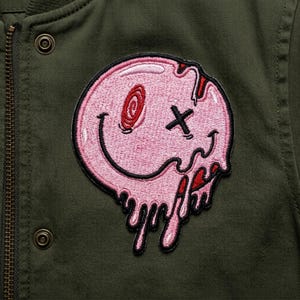 May include: A pink, melting smiley face patch with a black outline, a red spiral eye, and a black 'X' for an eye. The patch is on an olive green jacket.