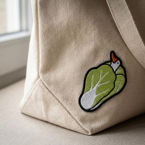 May include: Beige canvas tote bag with a green and white embroidered patch of a bird nestled in a leaf. The bag has a rectangular shape with a flat bottom and a long strap. The patch has a black border.