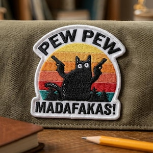 May include: Embroidered patch featuring a black cat holding two guns against a sunset backdrop. The patch reads "PEW PEW MADAF*KAS!" in white lettering. The patch is on a green canvas surface.