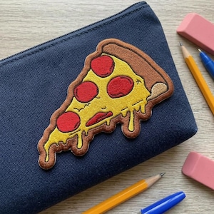 May include: A navy blue pencil case with a pizza slice patch. The patch features a yellow crust, red pepperoni, and melted cheese. Pencils, a pen, and an eraser are visible in the background.