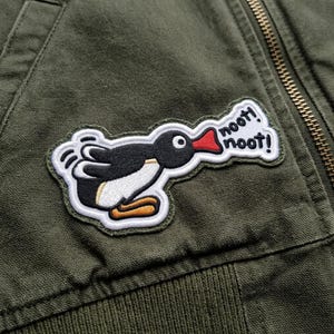 May include: Embroidered patch featuring a cartoon penguin with a black body, white belly, and orange feet. The penguin is saying "noot! noot!" in a speech bubble. The patch is on an olive green jacket.
