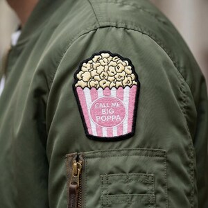Puede incluir: Chaqueta bomber verde oliva con un parche de palomitas de maíz. El parche presenta un recipiente de palomitas de maíz a rayas rosas y blancas con el texto "CALL ME BIG POPPA" en un óvalo blanco.