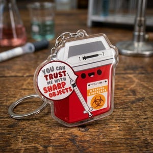 May include: A clear acrylic keychain featuring a red and white cartoon sharps container with a syringe graphic. The keychain has a speech bubble that reads "YOU CAN TRUST ME WITH SHARP OBJECTS". The container has a "WARNING BIOHAZARD" label.