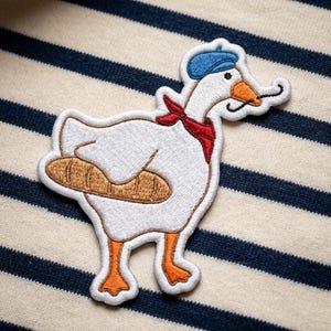 May include: Embroidered patch featuring a white goose wearing a blue beret, red scarf, and a pair of black mustaches. The goose is holding a brown baguette. The patch is on a striped background.