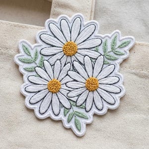 May include: Embroidered patch featuring three white daisies with yellow centers and green leaves. The patch has a scalloped edge and is set against a neutral background. The daisy design is a floral theme.