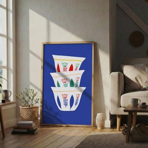 Op de afbeelding: Een ingelijste kunstdruk met een stapel witte koffiekopjes met bloemmotieven tegen een levendige blauwe achtergrond. De kopjes hebben rode, groene en gele accenten. Het kunstwerk wordt tentoongesteld in een woonkamer met natuurlijk licht.