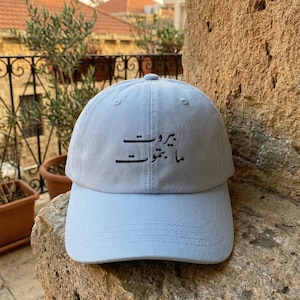 Puede incluir: Gorra de béisbol azul claro con texto árabe "Beirut Ma Btemout" bordado en la parte delantera. La gorra tiene una visera curva y un diseño clásico de seis paneles. El sombrero está hecho de una tela suave y duradera.