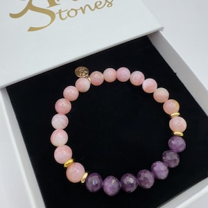 Pode incluir: Uma pulseira de contas com pedras preciosas rosa e roxas, acentuada com espaçadores dourados. A pulseira é exibida dentro de uma caixa branca com o texto "Angel Stones" e um logotipo dourado.
