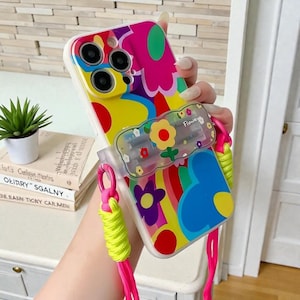 May include: A colorful phone case with a vibrant floral design in pink, yellow, blue, and purple. The case has a clear strap holder with the word "Flower" and a neon green and pink strap.