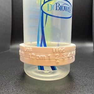 Könnte beinhalten: Ein hellbrauner 3D-gedruckter Babyflaschenring mit den Wochentagen und Zeiten aufgedruckt. Der Ring befindet sich um eine transparente Babyflasche mit blauen und weißen Akzenten. Der Text auf dem Ring enthält "Tues 12 pm 1 2 3 4" und "Wed".