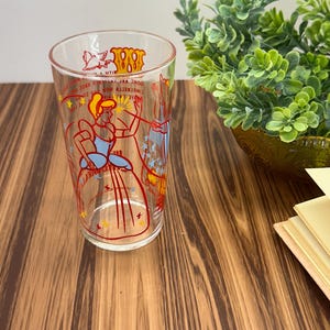May include: Clear glass tumbler featuring a Cinderella design in red, blue, and yellow. The illustration depicts Cinderella in her ball gown with text around the rim. A vintage collectible item.