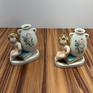 May include: Two ceramic figurines. Each features a cherubic figure kneeling beside a small vase. The vases are white with floral designs. The figures are on a stack of blue books, and the base is white.