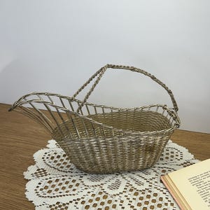 May include: A silver-colored metal basket with a handle, featuring a woven design and a unique shape. The basket sits on a white lace doily. A book with a pink cover is partially visible.