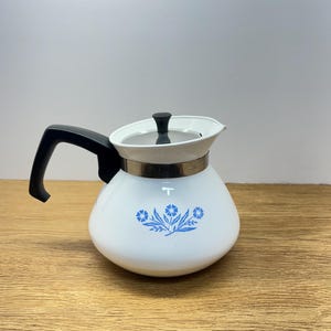May include: A white teapot with a rounded body and a black handle. The teapot features a blue floral pattern on the front, a silver-toned metal band around the neck, and a white lid with a small black knob handle on top.