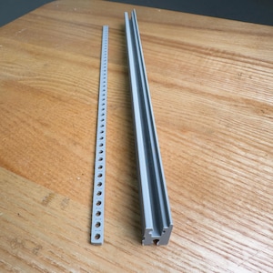 Könnte beinhalten: Zwei graue Kunststoffkomponenten auf einer Holzoberfläche. Eines ist ein langer, schmaler Streifen mit gleichmäßig verteilten Löchern. Das andere ist ein dickeres, gerilltes Teil, wahrscheinlich zur strukturellen Unterstützung oder Führung. Beide sind in einem neutralen Grauton gehalten.