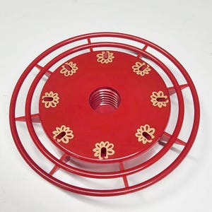 May include: A red hummingbird feeder with a circular design. The feeder has a central threaded port and a series of small, flower-shaped feeding ports. The outer ring is made of metal and has a cage-like structure.