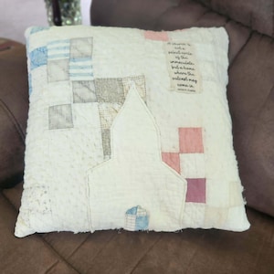 May include: A square quilted throw pillow featuring a central white fabric applique shaped like a church building. The background consists of a patchwork pattern in shades of cream, blue, and pink. A rectangular fabric label displays the text: A church is not a select circle of the immaculate but a home where the outcast may come in.