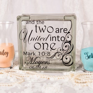 May include: A square glass block with black text reading and the two are united into one Mark 10:8 Moyers 06.09.18, flanked by two clear glass vases containing peach and turquoise sand, with the names Bailey and Seth printed on the respective vases.