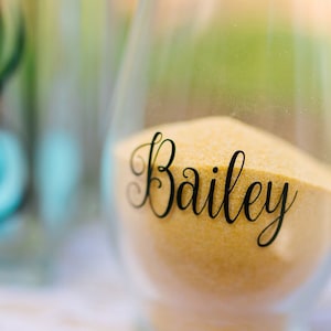 May include: A clear glass vessel filled with fine golden sand. The front of the glass features the name Bailey written in a black, elegant, cursive script. The container has a rounded, stemless shape and is partially filled with the sand.