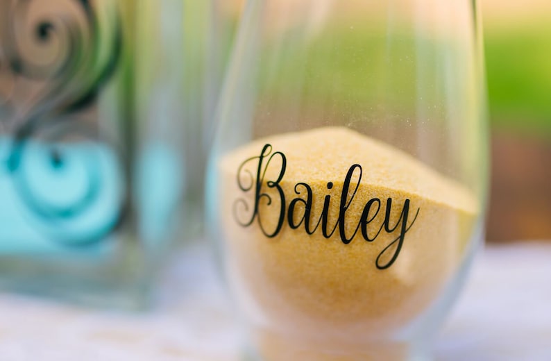 May include: A clear glass vessel filled with fine golden sand. The front of the glass features the name Bailey written in a black, elegant, cursive script. The container has a rounded, stemless shape and is partially filled with the sand.