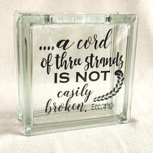 May include: A square, clear glass block featuring black lettering that reads, "....a cord of three strands IS NOT easily broken, Ecc. 4:12." The text is accompanied by a small, black, leafy vine graphic positioned to the right of the words.