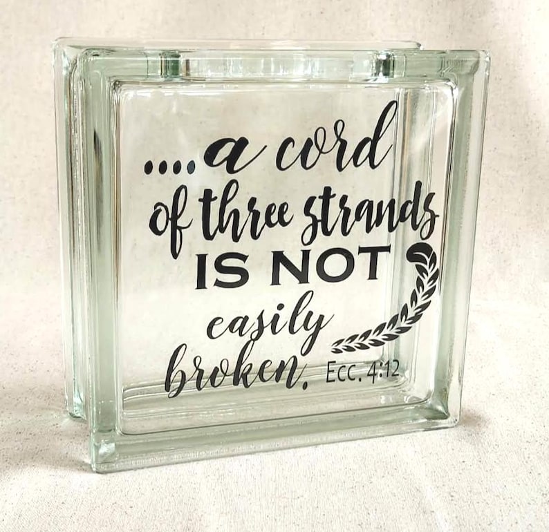 May include: A square, clear glass block featuring black lettering that reads, "....a cord of three strands IS NOT easily broken, Ecc. 4:12." The text is accompanied by a small, black, leafy vine graphic positioned to the right of the words.
