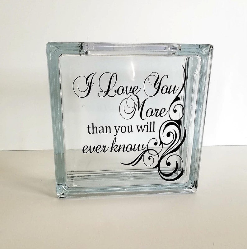 May include: A square glass block with a hollow center and a slot at the top. The front surface features black cursive lettering that reads, I Love You More than you will ever know, accompanied by a decorative black swirl design on the right side.