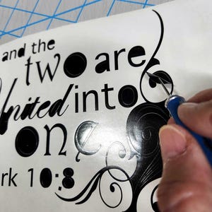 May include: A black vinyl decal featuring the text and the two are united into one and the reference Mark 10:8, accompanied by decorative swirling flourishes. A blue-handled weeding tool is shown removing excess vinyl from the design.