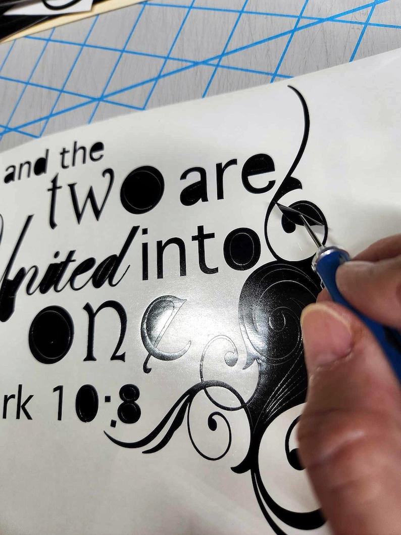 May include: A black vinyl decal featuring the text and the two are united into one and the reference Mark 10:8, accompanied by decorative swirling flourishes. A blue-handled weeding tool is shown removing excess vinyl from the design.