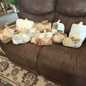 May include: A collection of ten fabric pumpkins in various shapes and sizes, featuring assorted patchwork patterns, quilted textures, and neutral color palettes including cream, beige, brown, and muted green, each topped with a small fabric stem.