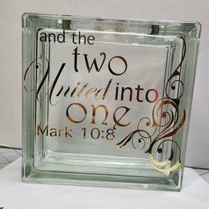 May include: A square, clear glass block featuring gold metallic lettering that reads, and the two united into one, Mark 10:8. The text is accompanied by decorative gold filigree swirls on the right side of the block, creating a reflective surface.