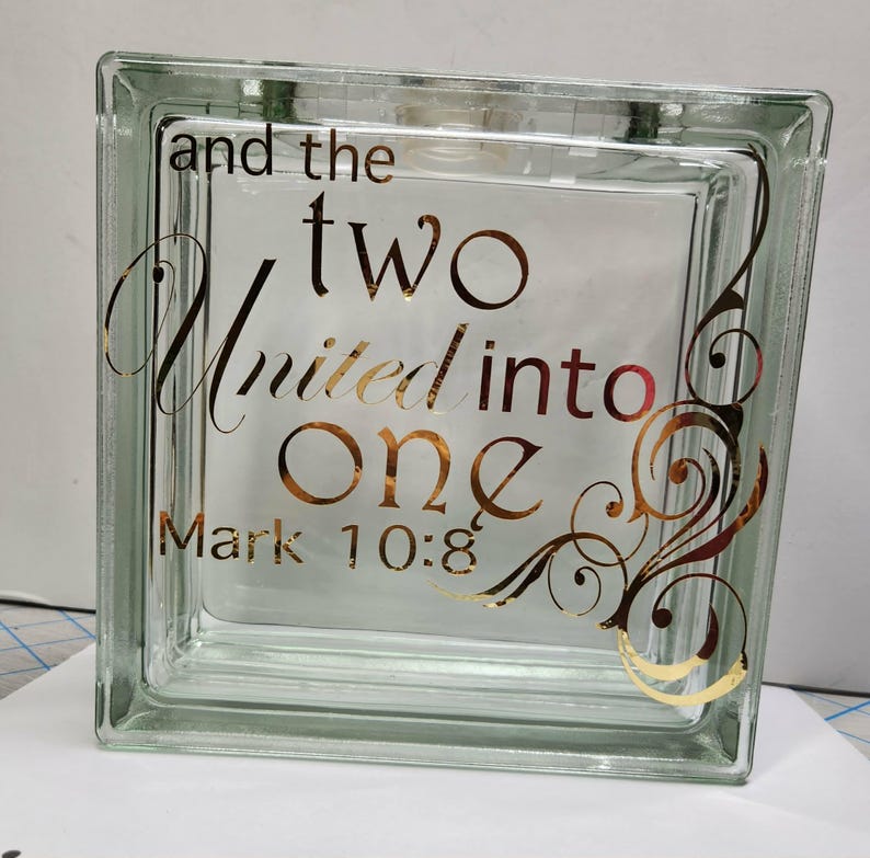 May include: A square, clear glass block featuring gold metallic lettering that reads, and the two united into one, Mark 10:8. The text is accompanied by decorative gold filigree swirls on the right side of the block, creating a reflective surface.