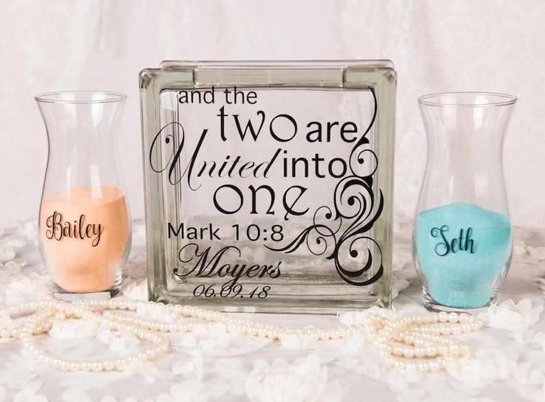 May include: A square glass block with black text reading and the two are united into one Mark 10:8 Moyers 06.09.18, flanked by two clear glass vases containing peach and turquoise sand, with the names Bailey and Seth printed on the respective vases.