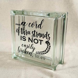 May include: A square, clear glass block featuring black lettering that reads, "...a cord of three strands IS NOT easily broken. Ecc. 4:12," accompanied by a decorative leaf-like flourish at the end of the text, creating a transparent display piece.